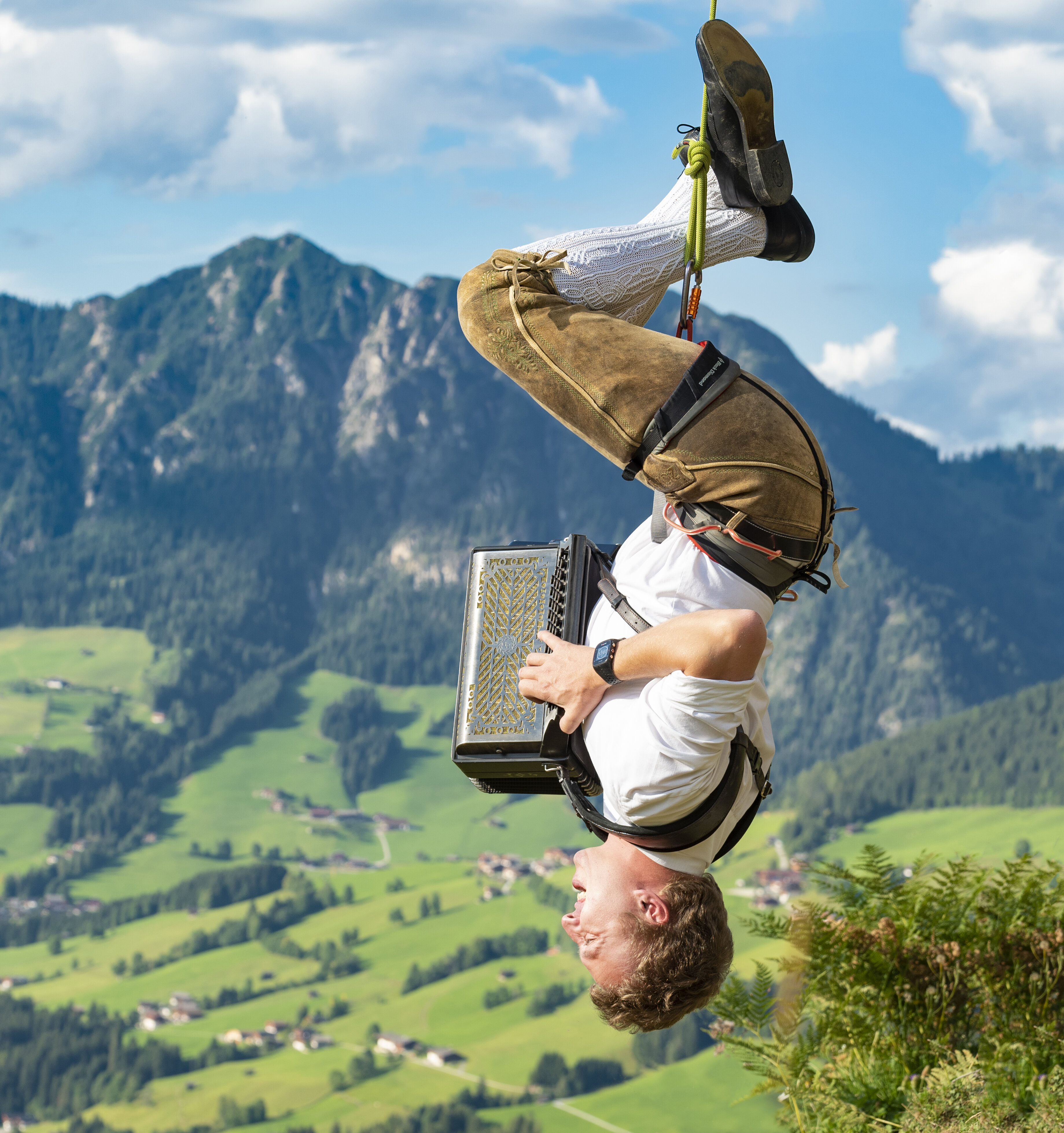 Ein Mann hängt an einem Seil kopfüber, in Lederhose und weißem Hemd spielt er mit seiner Ziehharmonika | © Alpbacher Plattler